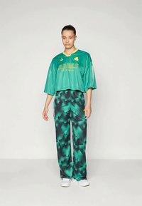 Haut court surdimensionné vert à manches courtes avec logo jaune, associé à un pantalon vert à motifs comportant des nuances de vert et de noir. Chaussures blanches.