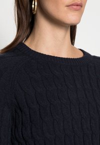 Maglione lavorato a maglia blu navy con motivo a trecce, caratterizzato da scollo rotondo e polsini a costine. Texture morbida e peso medio.