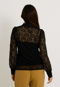 Blusa de encaje negro con patrones texturizados y mangas largas, con cuello alto y tela semitransparente, combinada con pantalones de color mostaza.