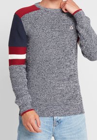 Pull en maille avec un corps texturé bleu marine, des manches raglan rouges et bleues, et des rayures blanches sur le bras rouge. Encolure ronde. Coupe décontractée.