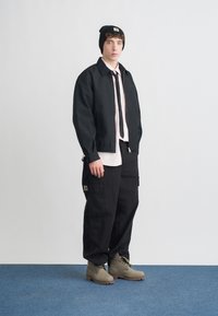 Μαύρο σακάκι με φερμουάρ στο μπροστινό μέρος, ροζ πουκάμισο, μαύρη γραβάτα, παντελόνι cargo και γκρι μπότες. Μαύρος σκούφος με λογότυπο. Φόντο μπλε μοκέτας.