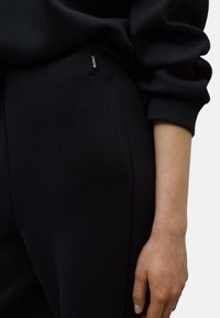 Sudadera negra de corte relajado, con una etiqueta de logo visible en el lateral. Tejido de algodón suave con un aspecto ligeramente texturizado.