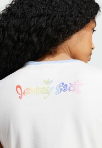 Biela tričko s krátkym rukávom a modrým golierom, s viacfarebným textom 'Jeremy Scott' a logom Adidas na chrbte. Hladká textúra.