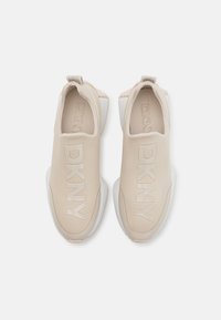 Zapatos slip-on beige con la marca DKNY, parte superior de sintético suave, cuello acolchado y suela de goma blanca. Diseño ligero y minimalista.