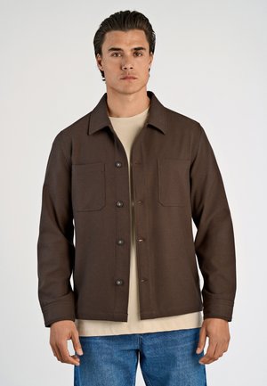 Mand iført en brun knappet jakke over en beige skjorte og blå jeans, stående mod en ensfarvet lys baggrund.
