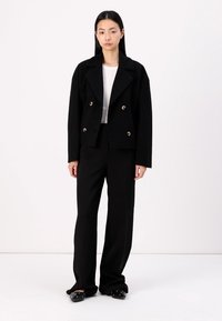 Black double-breasted jacket with large metallic buttons, textured fabric, and wide lapels, paired with wide-leg black trousers and flat shoes.