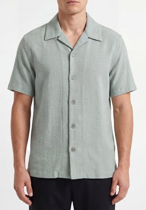 Hombre con camisa gris claro de manga corta con botones y cuello campestre, y pantalones negros, de pie sobre un fondo blanco.