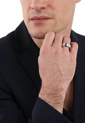 Hombre con chaqueta negra tocándose la barbilla, lleva un anillo plateado en el dedo medio con una inscripción grabada.