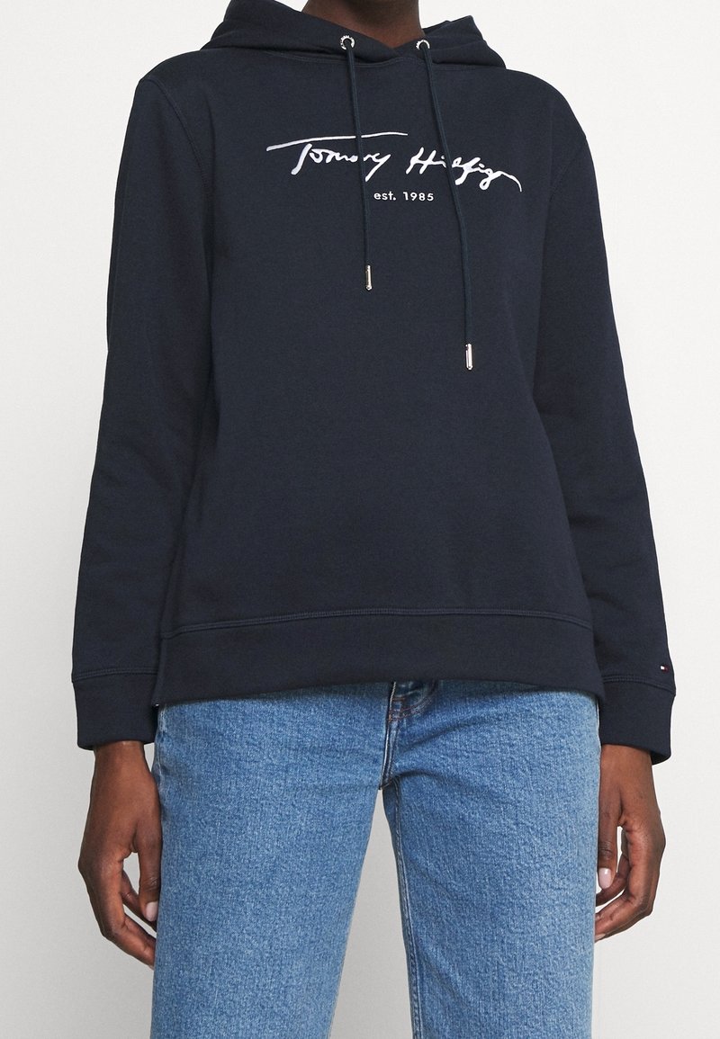 Personne portant un sweat à capuche bleu marine Tommy Hilfiger avec cordons et texte "est. 1985", associé à un jean bleu clair.
