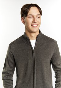 Grijze zip-up trui met ribbelstructuur, hoge kraag en lange mouwen. Model draagt een wit T-shirt eronder en lacht tegen een lichte achtergrond.