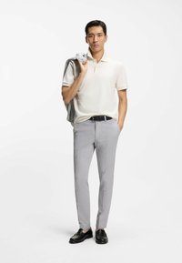 Vaaleanbeige pikeepaita, harmaat räätälöidyt housut, mustat kengät. Vaaleanharmaa bleiseri roikkuu yhdellä olkapäällä. Sileät tekstuurit, minimalistinen muotoilu.