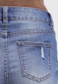 Jeans de mezclilla azul con un bolsillo trasero desgastado que presenta bordes deshilachados. Corte estándar con costuras básicas y una cinturilla de altura media.