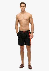 Pantaloni da nuoto neri con logo arancione "SUPERDRY", dotati di vita con cordino, lunghezza sopra al ginocchio e una texture di tessuto liscia.