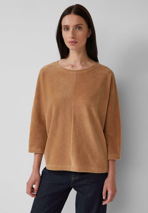 Femme portant un haut léger et texturé de couleur marron clair à manches longues et un jean bleu foncé, se tenant contre un fond gris uni.