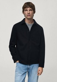 Giacca camicia in misto lana nera con colletto, bottoni frontali e tasca sul petto, abbinata a jeans in denim azzurro chiaro.