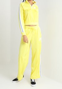 Gul zip-up sweatshirt med långa ärmar och vita ränder, parat med matchande gula sweatpants med dragsko och logotyp.