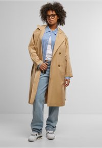 Tan trench coat, double-breasted design, light blue striped shirt underneath, relaxed-fit jeans, white sneakers with black accents.