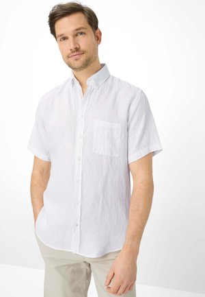 Mand med kort brunt hår iført en hvid, kortærmet skjorte med knapper, stående med den ene hånd i lommen mod en hvid baggrund.