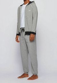 Giacca grigia con cappuccio e dettagli neri, tasche con zip e logo; abbinata a pantaloni della tuta grigi coordinati con vita elastica.