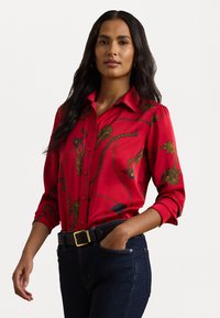 Red blouse with a collar, featuring gold and blue patterns of ropes and medallions. Worn with dark denim jeans and a black belt.