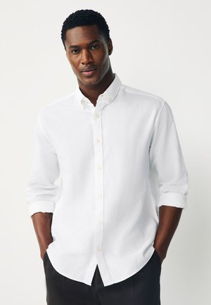 Mand iført en hvid skjorte med knapper og opkørte ærmer og sorte bukser, stående med hænderne i lommerne mod en ensfarvet baggrund.