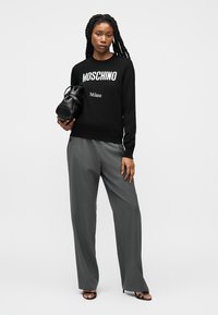 Femme aux longues tresses portant un sweat-shirt noir Moschino Milano, un pantalon gris à jambes larges, des sandales noires et tenant un sac à main noir.