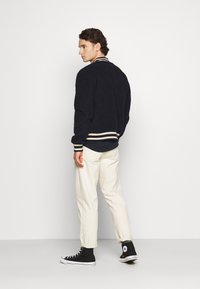 Giacca bomber in pile blu marino con polsini e colletto a strisce beige, abbinata a pantaloni beige chiaro e sneaker alte nere.