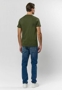 Olivgrön kortärmad t-shirt i bomull, rund halsringning, matchad med blå denimjeans och vita sneakers. Synlig detalj vid bakre halsringning.