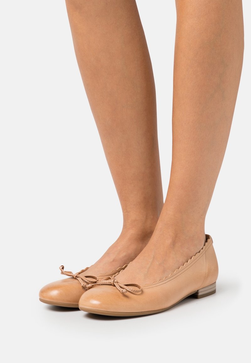 Pieds chaussés de ballerines en cuir beige avec des bords ondulés et de petits nœuds décoratifs, présentés sur un fond clair uni.
