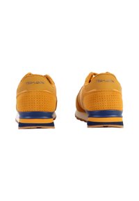 Zapatillas deportivas amarillas con acabado de ante, laterales perforados, acentos de rayas azules y amarillas en la suela, y detalle de logo en el talón.