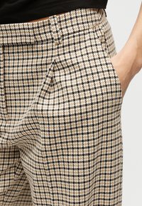 Μπεζ, μαύρο και άσπρο καρό παντελόνι με ένα χέρι στην δεξιά τσέπη, φορεμένο με μαύρο τοπ.