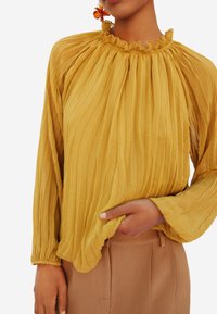 Blusa plissettata giallo senape con collo arricciato e maniche lunghe, abbinata a pantaloni beige. Le pieghe creano texture e movimento.