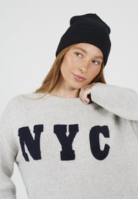 Młoda kobieta w czarnej dzianinowej czapce i jasnoszarym swetrze z dużymi czarnymi literami "NYC," patrząca w bok z ręką blisko twarzy.