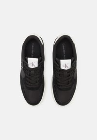 Černé sportovní boty s kombinací hladkých a strukturovaných materiálů, s černými šňůrkami, bílým logem a bílou podrážkou.