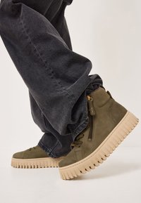 Baskets montantes vert olive avec une finition en daim texturé, dotées d'une épaisse semelle en caoutchouc crème et d'un détail de fermeture éclair sur le côté.