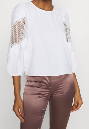 Vrouw die een witte blouse met kanten inzetmouwen en mauve satijnen op maat gemaakte broek draagt, geshowd van nek tot dijen tegen een effen achtergrond.