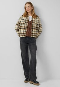 Karierte Jacke in Creme, Braun und Schwarz mit kastiger Passform; kombiniert mit einer braunen Strickjacke, schwarzen Jeans und klobigen schwarzen Stiefeln.