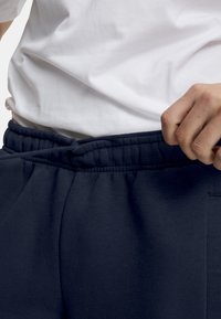 Mørkeblå joggebukser med elastisk linning og snøring, med en myk, glatt tekstur og sidelommer. Brukt sammen med en hvit t-skjorte.