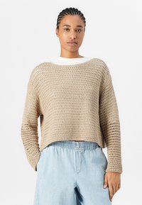 Beige Häkelpullover mit weitem Halsausschnitt und langen Ärmeln, der eine offene Strickstruktur aufweist. Kombiniert mit hellblauen, locker sitzenden Hosen.