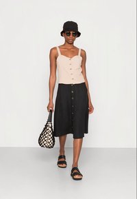Top cropped beige com botões, saia preta em linha A com botões frontais, chapéu preto estilo bucket, óculos de sol redondos e uma bolsa preta em tecido trançado. Sandálias rasas.