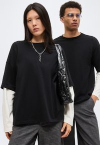 Νέα γυναίκα και άνδρας με στρωτά μαύρα και λευκά τοπ και γκρίζες καρό παντελόνες, η γυναίκα κρατάει γυαλιστερή μαύρη τσάντα ώμου και φοράει ασημένιο αλυσίδωτο κολιέ.