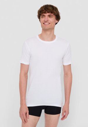 White short-sleeve t-shirt made of smooth fabric with a crew neckline, worn with black fitted shorts. Model is standing with a neutral expression.
