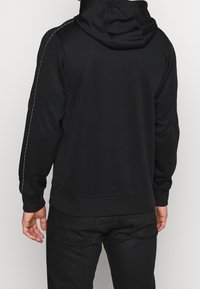 Schwarzer Hoodie mit Kängurutasche, Kapuze mit Kordelzug und kontrastierenden weißen Nähten an den Ärmeln. Gerippte Bündchen und Saum.