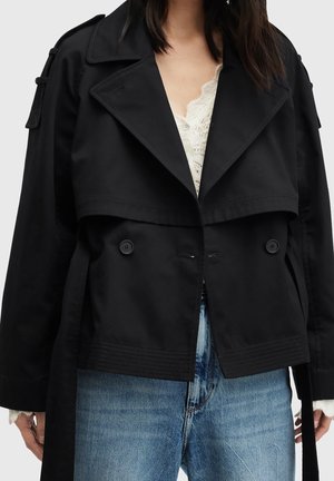 Femme portant un trench-coat court noir par-dessus un haut en dentelle blanche et un jean bleu, avec le manteau partiellement boutonné et la ceinture pendue.