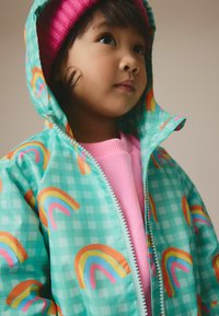Veste de pluie pour enfant en bleu clair, dotée d'un motif à carreaux et d'impressions colorées de rainbow. Fermeture éclair, portée par-dessus un pull rose.