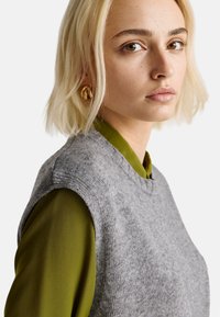 Šedá pletená vesta s kulatým výstřihem, přeložená přes zelenou košili s dlouhým rukávem. Měkká textura a čisté linie, minimální detaily.