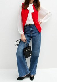 Rote ärmellose Weste über einer weißen Bluse, kombiniert mit weit geschnittenen blauen Jeans. Accessoire: schwarze gesteppte Handtasche mit goldenen Details.