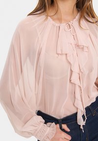Blusa in chiffon rosa chiaro con scollo arricciato, parte anteriore con volant e maniche a sbuffo. Presenta polsini elasticizzati e una vestibilità ampia.