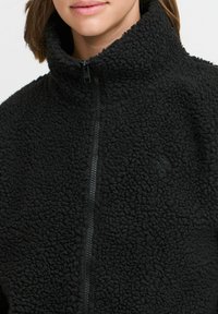 Veste en polaire noire avec un col haut, fermeture éclair et tissu texturé. Elle présente un petit logo sur la poitrine.