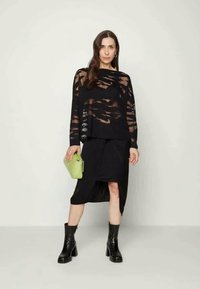 Camisola preta de mangas longas com padrões recortados, combinada com uma saia assimétrica preta. Acessórios incluem uma mala verde e botas de tornozelo pretas.